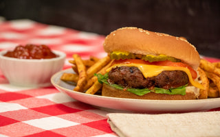 a thick Halal burger patty with a sesame bun, pickles, tomatoes, and cheese on top and lettuce under it  sits on a plate with fries around it. 