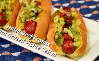 Three beef franks are lined up on a white plate. On each of the franks is pickle relish with large chunks of pickles 
