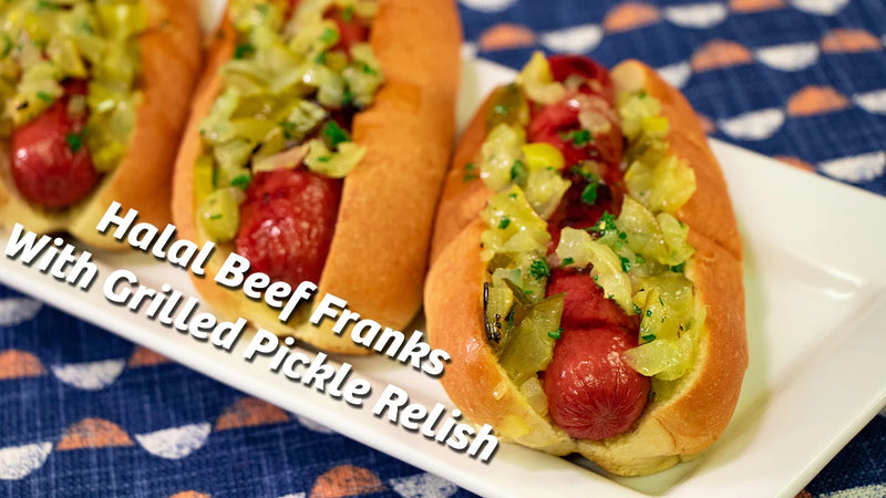 Three beef franks are lined up on a white plate. On each of the franks is pickle relish with large chunks of pickles 