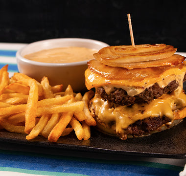 A double cheeseburger with two beef patties and cheese between slices of fried onion, served with fries and a white bowl of sauce. All served on a black plate on a white and blue striped tablecloth.