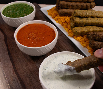 Three white bowls of green, red, and white kebab sauces on a black serving board. Behind the board is a rectangular white plate with yellow rice underneath an assortment of kebabs. 