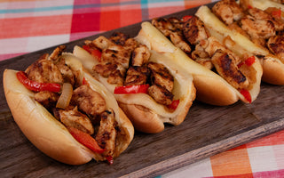 An assortment of chicken sandwiches with chunks of chicken, sliced red peppers and onion, and melted cheese on hotdog buns. All served on a black serving board on a vibrant background.