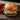 A Midamar seasoned beef patty rests on a bed of lettuce, tomato, red onion, and american cheese. The burger is served on a black wooden board and has fried onion rings beside it